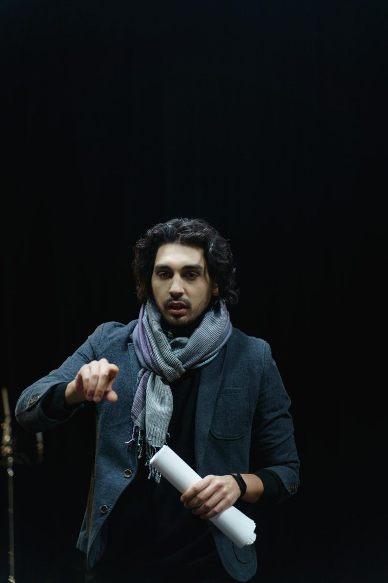 A male director in a dark theater, guiding rehearsals while holding a script.