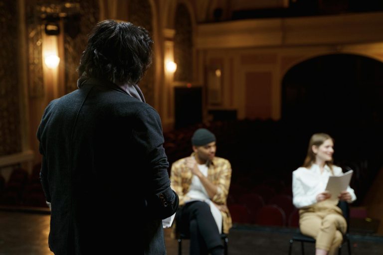 Actors rehearse on a classical theater stage with scripts before performance.